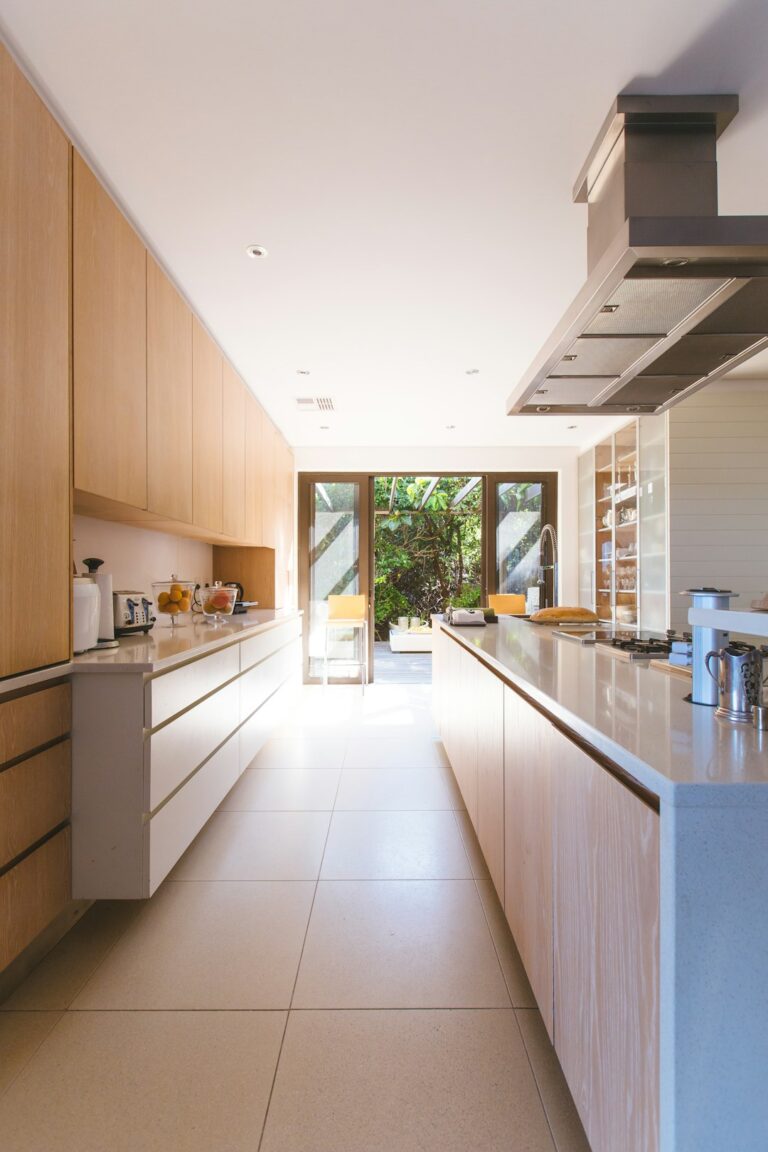 A bright kitchen with natural stone countertops, wood cabinets, and sunlight streaming through large windows.